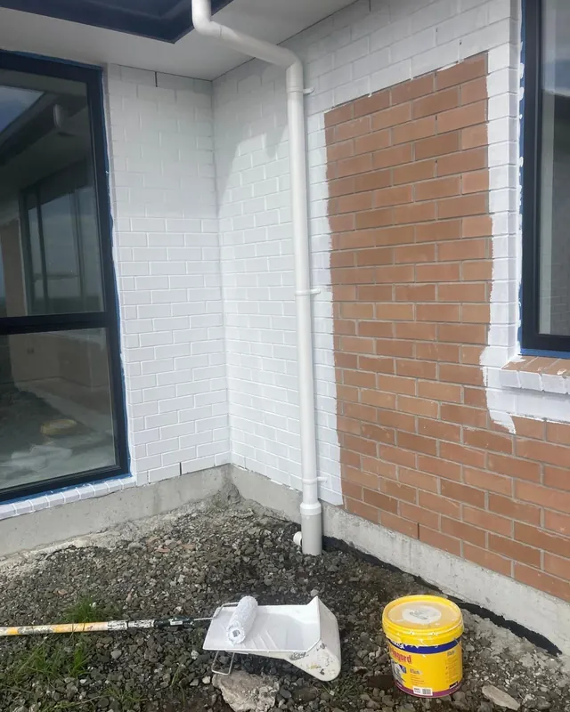 Brick wall half painted white, with a roller, tray and paint bucket on the ground
